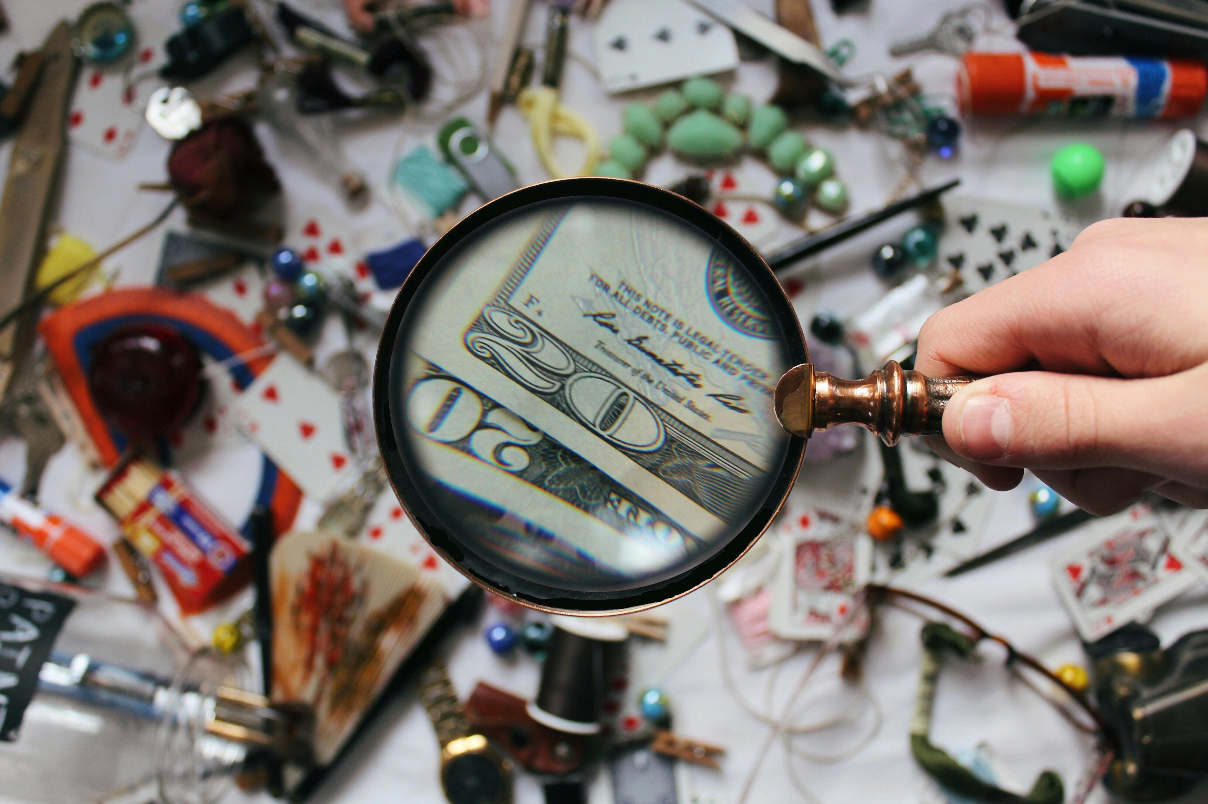 Hand holding a magnifying glass over a cluttered desk, highlighting the intricate details on a five-dollar bill, illustrating the concept of close examination similar to how to analyze Python classes with the inspect module.