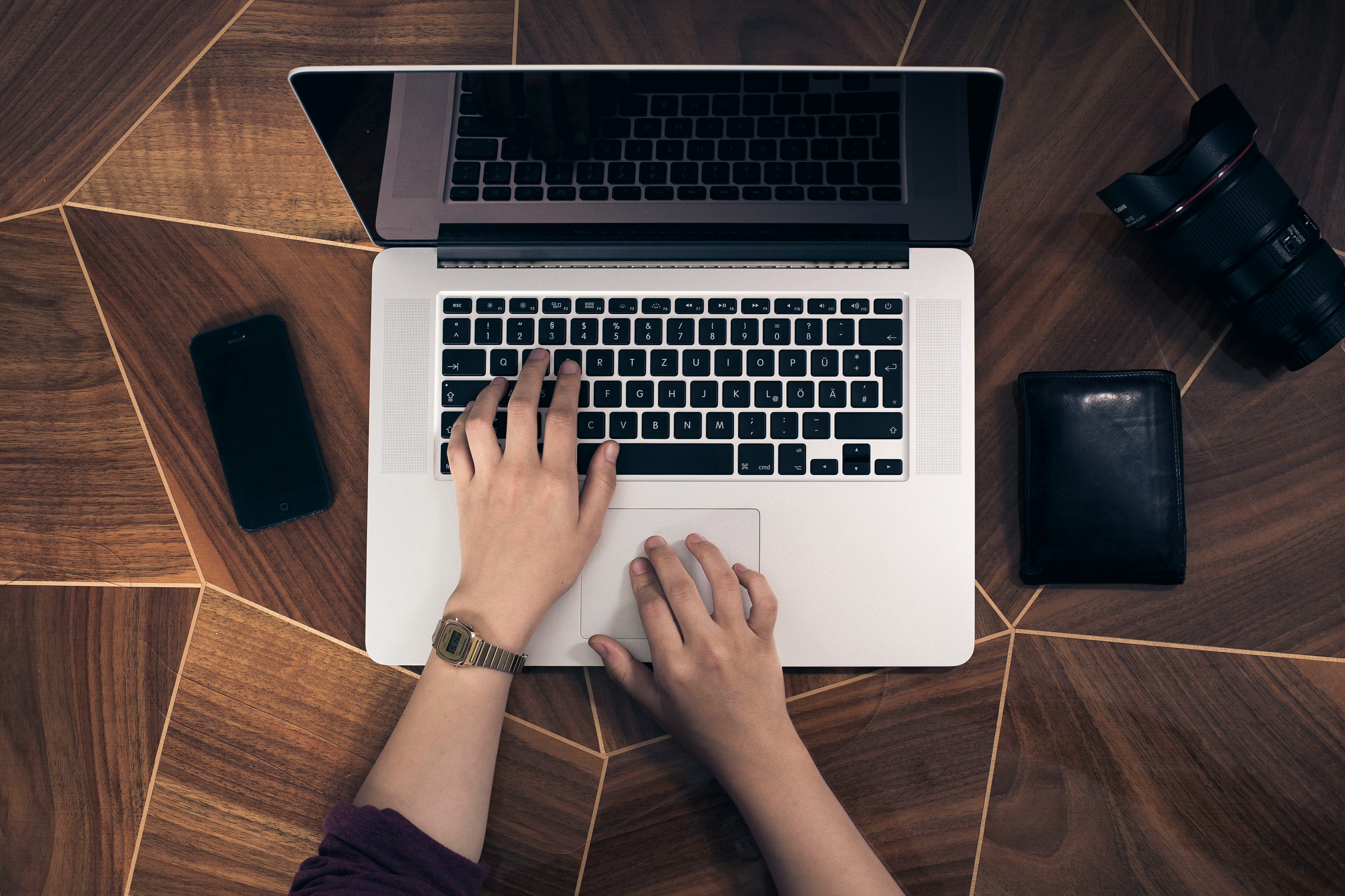 Person typing on a laptop, learning Python 3.12 generics syntax for improved type-hinting and cleaner code. Nearby items include a smartphone, camera lens, and wallet on a geometric wooden table.